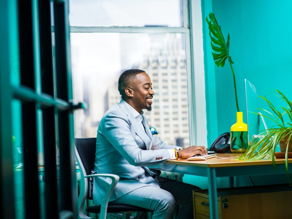 Um homem negro, vestindo terno cinza, está sentado em um escritório, mexendo em um computador. Ele sorri como se estivesse relaxado. É possível ver algumas plantas no ambiente. Otimizar processos internos
