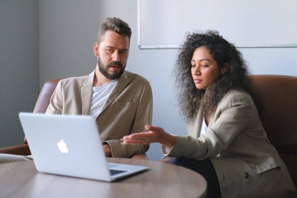 Um homem branco e uma mulher negra estão sentados em um escritório. Eles conversam sobre algo, enquanto a mulher aponta para a tela de um laptop prateado, que está em cima de uma mesa de madeira.