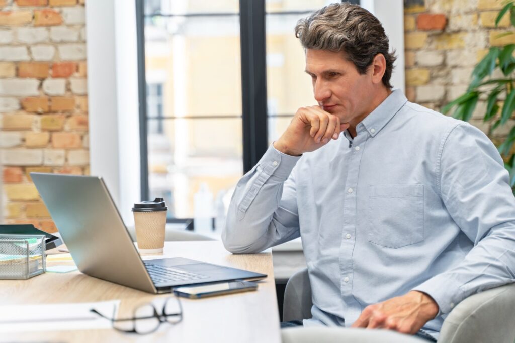 Um homem branco de cabelo curto e grisalho, vestindo camisa social azul-claro, está sentado em um escritório, de frente para um laptop prateado. O dispositivo está em cima de uma mesa de madeira, na qual também estão um copo de café, um celular, óculos e alguns papéis.
