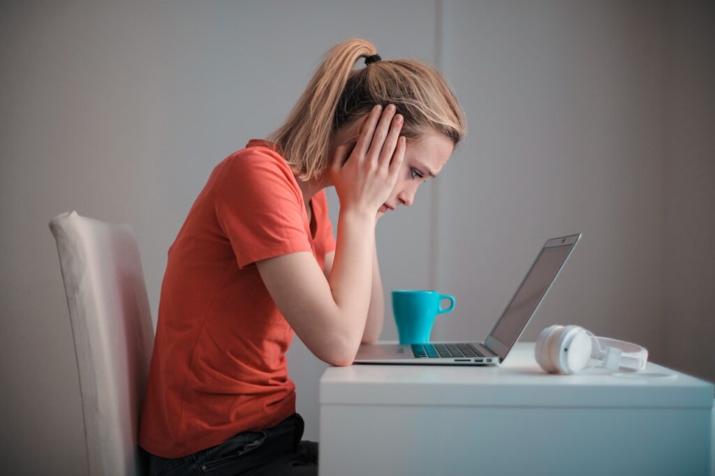 Uma mulher branca, com cabelo loiro e rabo de cavalo, vestindo uma camiseta laranja, está sentada em frente a um laptop. Ela está com as mãos no rosto, demonstrando sinais de preocupação com o fim da EIRELI.