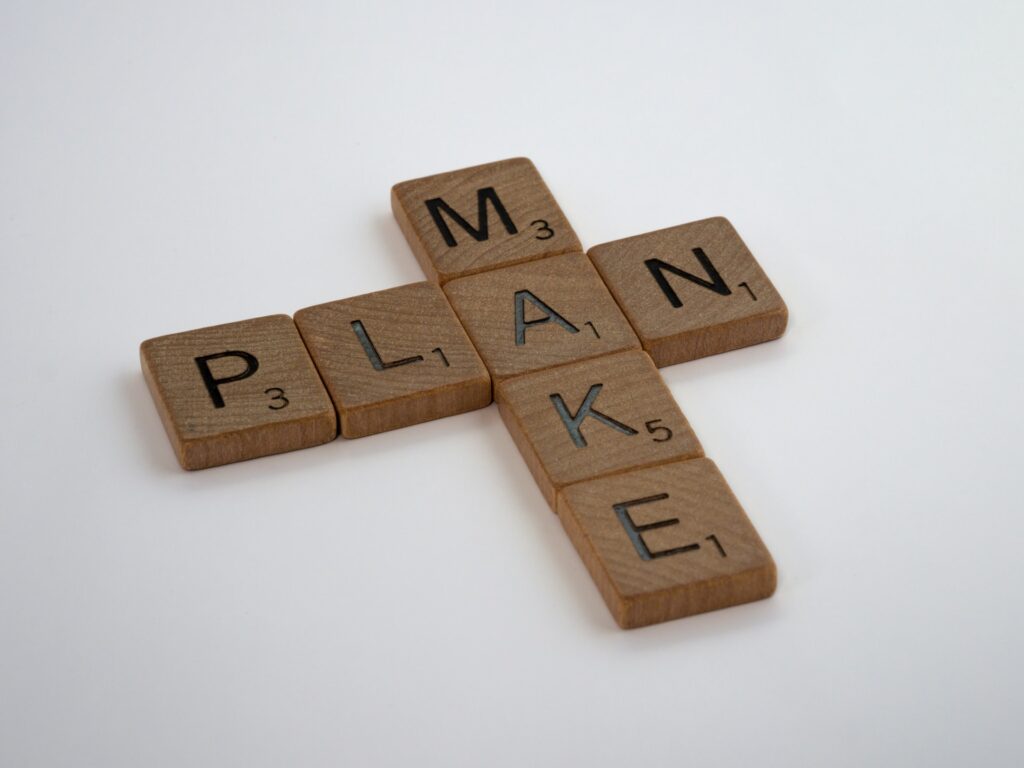 Sete peças de madeira quadradas com letras, organizadas de maneira cruzada, formando as palavras “Make” e “Plan”, “Fazer” e “Planejar” em inglês. O cruzamento acontece na letra A.
