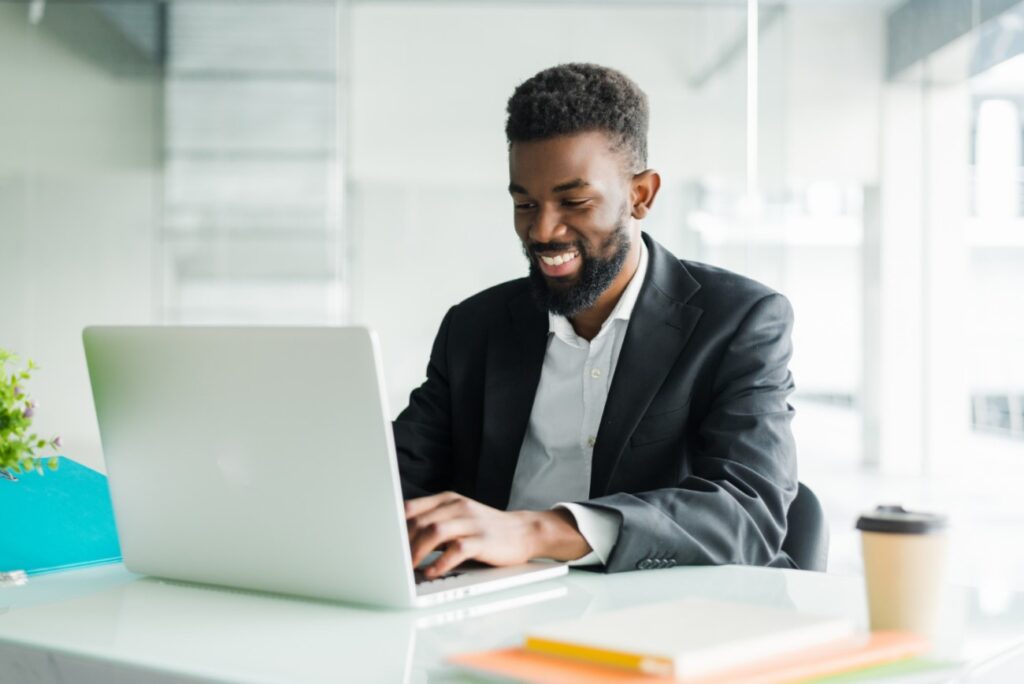 Um homem negro, vestindo terno, está sentado em uma mesa de escritório, mexendo em um laptop. Ele está sorrindo, simbolizando os resultados positivos de seu negócio.