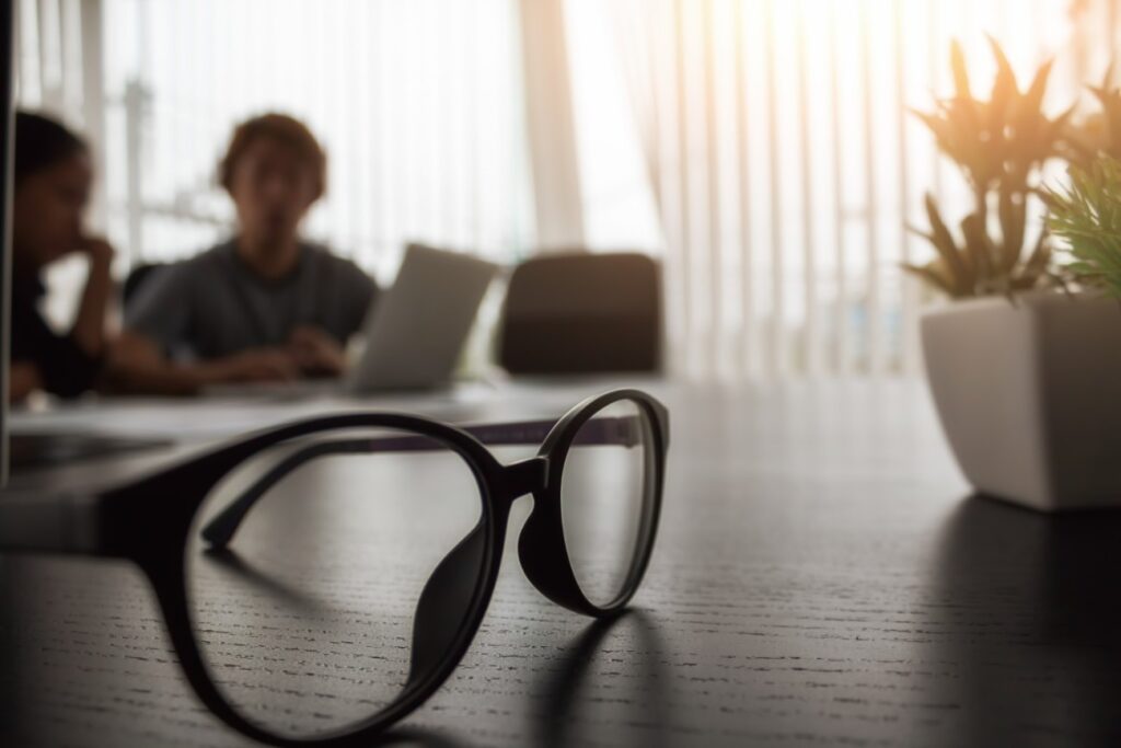 o que é escrituração contábil. Óculos de grau sobre uma mesa de madeira de um escritório, com um vaso de planta ao lado. Ao fundo, fora de foco, duas pessoas sentadas, mexendo em um laptop.
