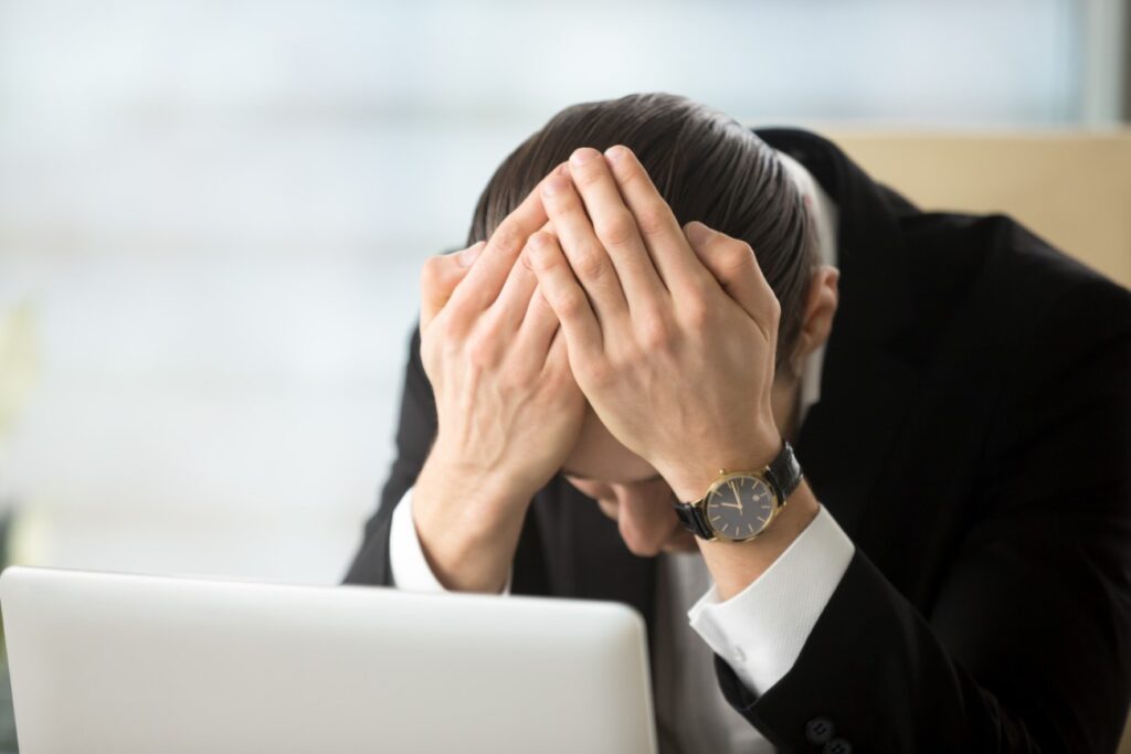 Um homem branco, vestindo terno preto e usando um relógio de pulso, está sentado em um escritório com as mãos na cabeça, como se estivesse preocupado. Há um laptop prateado à sua frente.