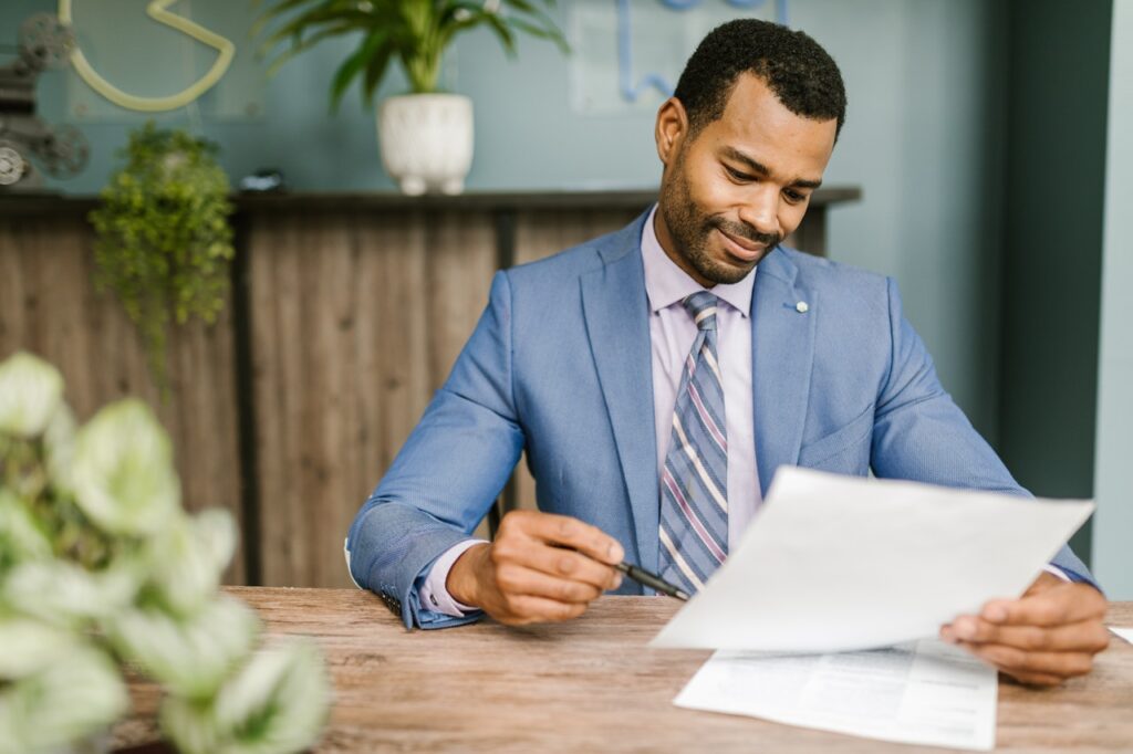 Um homem negro, vestindo terno azul, está sentado em um escritório. Ele analisa alguns documentos. Toda a mobília do ambiente é de madeira e há várias plantas espalhadas pelo lugar.