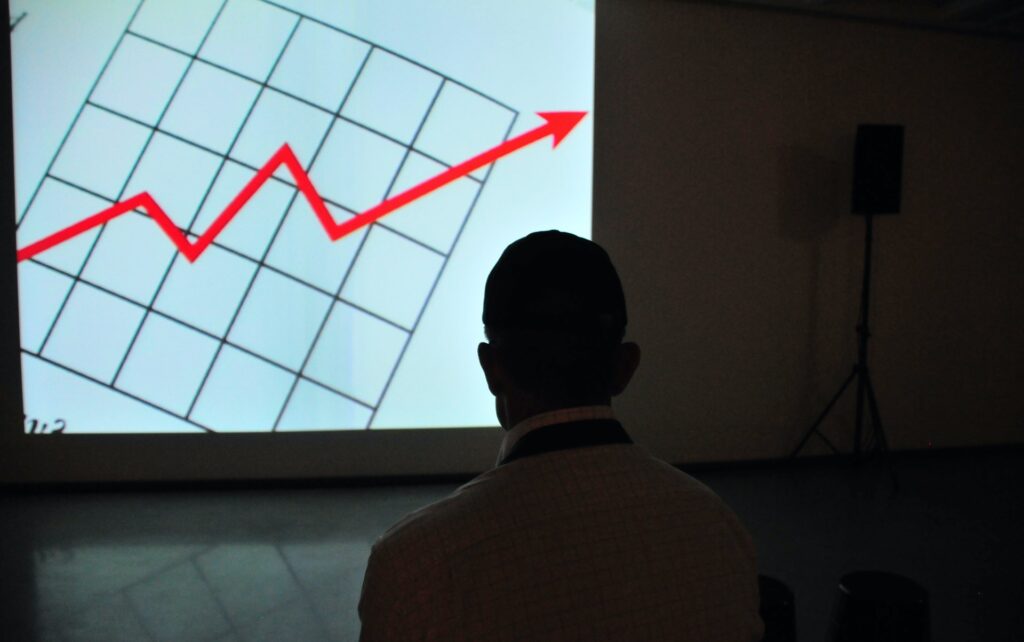 Um homem de costas em uma sala escura, olhando para uma projeção na parede. Na imagem do projetor, vemos um grid com uma seta vermelha oscilante, simbolizando um gráfico de desempenho.