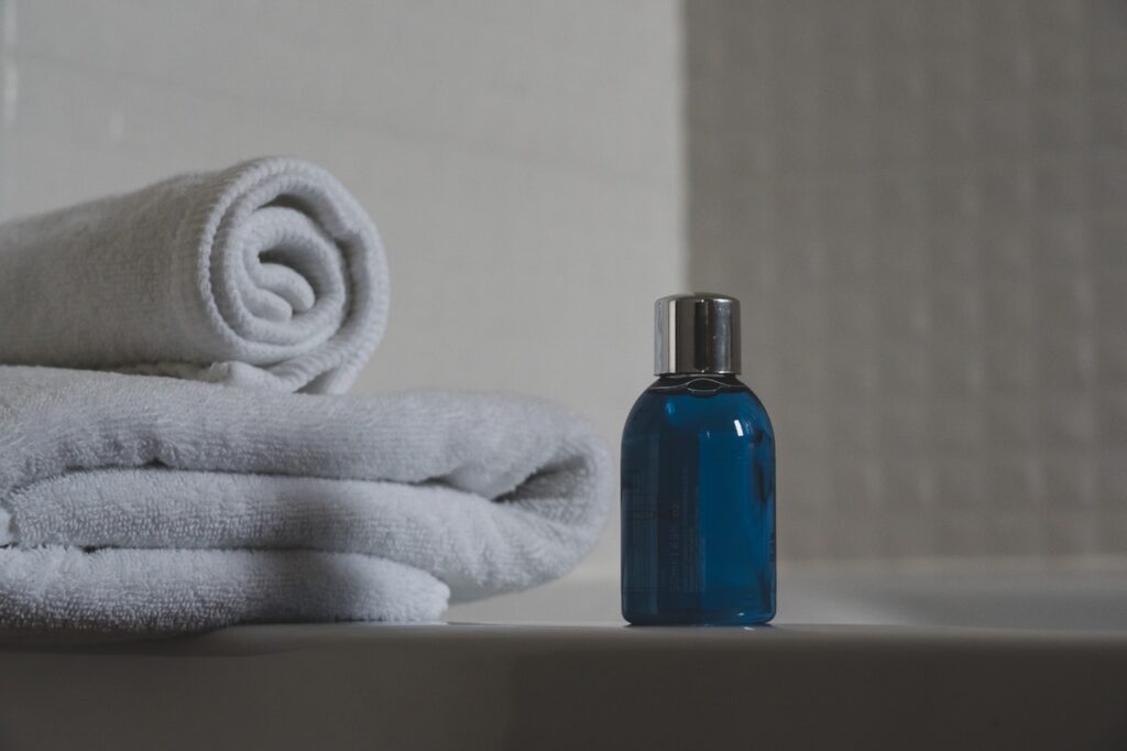 Duas toalhas, uma dobrada e outra enrolada, estão na borda de uma banheira. Um frasco azul está ao lado. Tanto a banheira quanto os revestimentos são brancos.