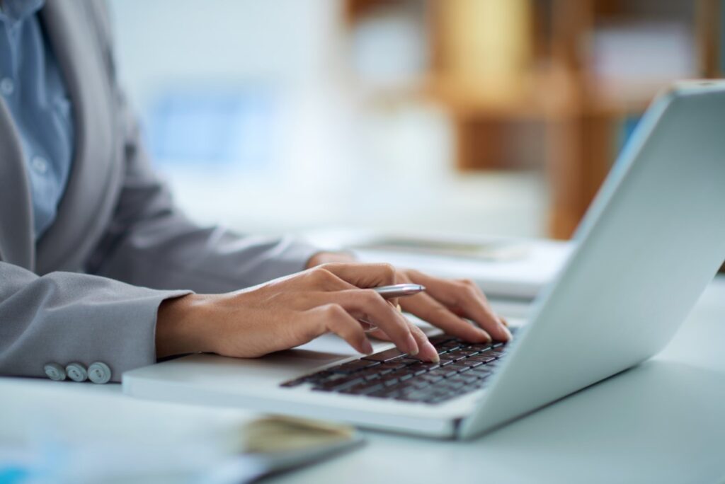 Uma pessoa branca, vestindo terno cinza e camiseta social azul-claro, está mexendo em um laptop branco, enquanto segura uma caneta prateada.