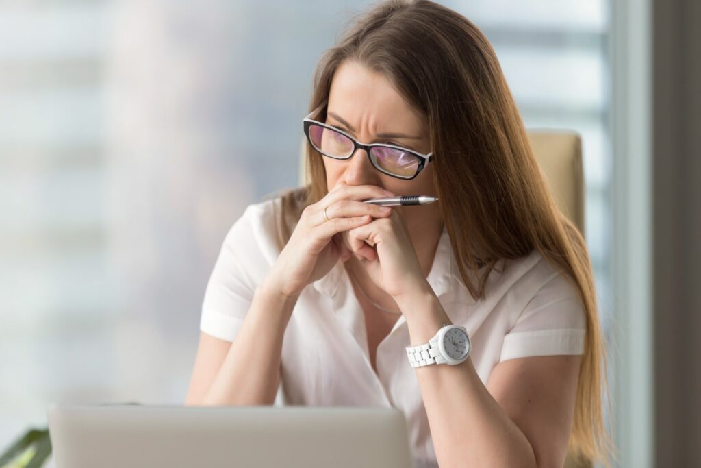 Uma mulher branca, vestindo camiseta também branca, usando um relógio de pulso branco e óculos nas cores branco e preto, está sentada em um escritório, em frente a um computador. Ela está com as mãos no rosto, indicando preocupação.