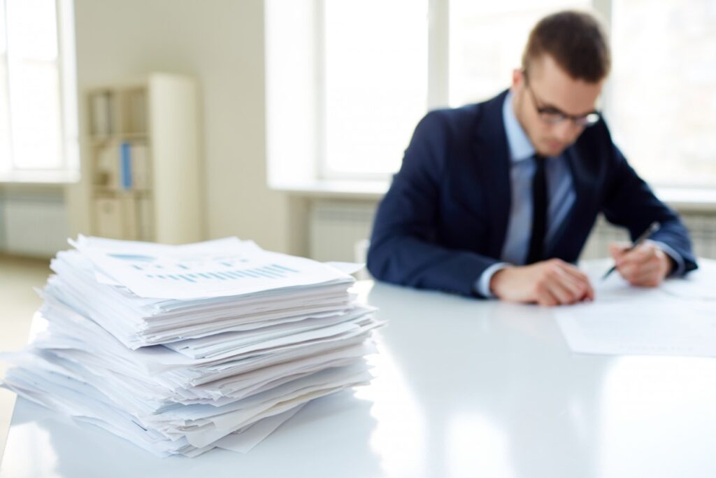 Um homem branco, de cabelo curto, vestindo terno e usando óculos, está sentado em uma mesa, preenchendo alguns documentos. À sua frente, em primeiro plano, está uma pilha de outros documentos.