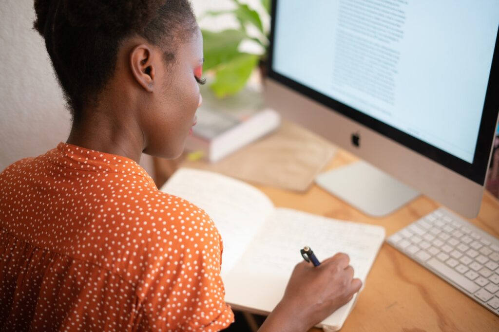 Uma mulher negra, vestindo camiseta laranja cheia de bolinhas brancas, está sentada em uma mesa de escritório, de frente para um computador, fazendo anotações em um caderno.