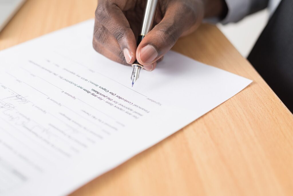 Uma pessoa negra assinando um contrato que está sobre uma mesa de madeira.