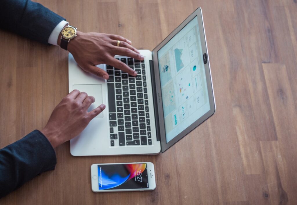 Um homem negro, usando terno, relógio de pulso e aliança de casamento, está mexendo em um laptop prateado. Na tela do aparelho há vários gráficos de desempenho. Ao lado do laptop, apoiado sobre uma mesa de madeira, está um celular. Apenas os braços do homem estão visíveis.