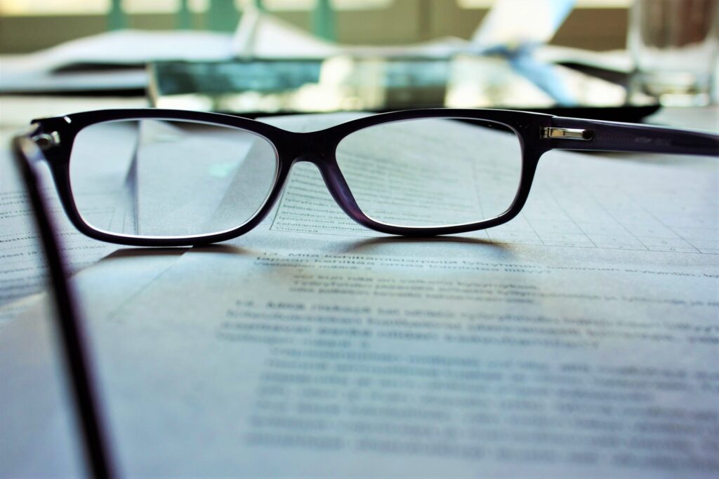 Um par de óculos sobre vários documentos em cima de uma mesa. Ao fundo, há vários outros objetos desfocados.