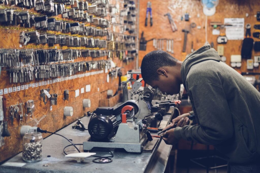 Um homem negro, vestindo blusa de moletom cinza-escuro, está em uma oficina, fabricando chaves. À sua frente, há uma bancada repleta de ferramentas e um painel na parede cheio de chaves penduradas.