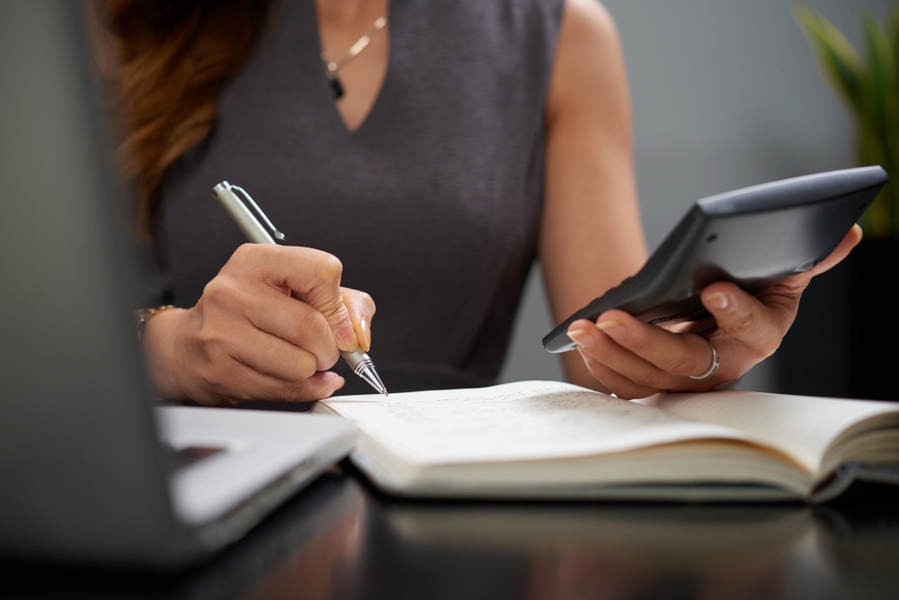 Vemos uma mulher sentada à mesa, fazendo anotações em um caderno enquanto faz cálculos em uma calculadora. Em sua frente, à um notebook aberto, para ilustrar a importância da contabilidade para a gestão empresarial.