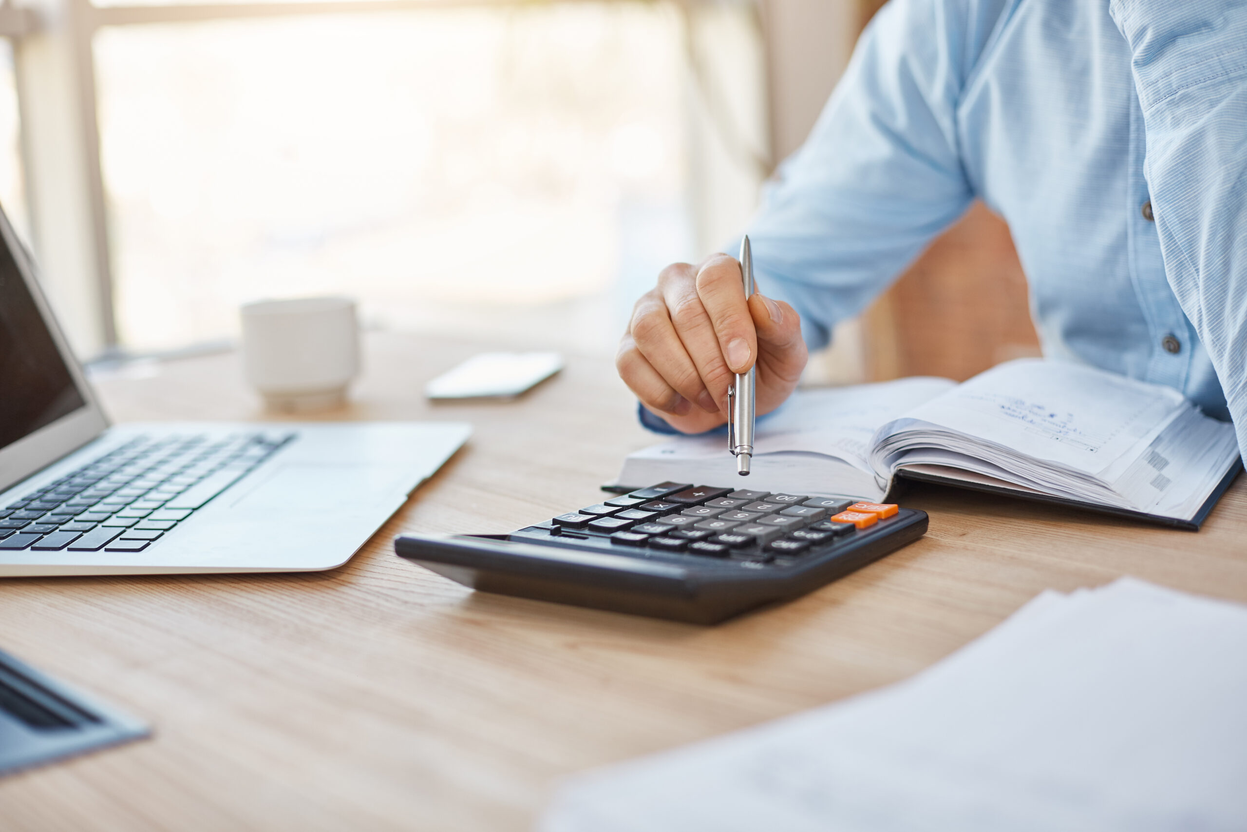 Vemos o braço de um homem, vestindo uma camisa azul clara, mexendo em uma calculadora com uma caneta. Em sua frente, também vemos um caderno e, mais ao lago, um notebook aberto e uma xícara de café. Tudo isso para ilustrar o artigo sobre mudanças do Simples Nacional em 2023