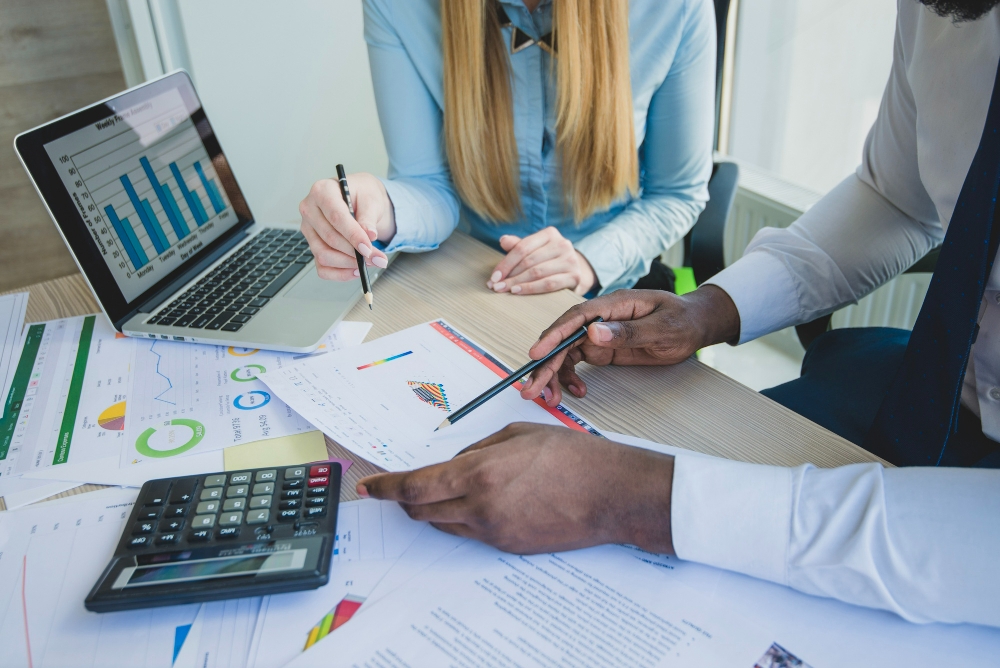 Homem e mulher em mesa de escritório, com gráficos, computador e calculadora, discutindo as contabilidades de uma empresa, para ilustrar artigo sobre as principais responsabilidades de uma consultoria contábil