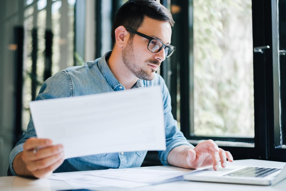 Foto contador de negócios casual sério, pensativo, focado e jovem, no escritório, olhando e trabalhando com papéis e documentos de declaração de imposto de renda e laptop para ilustrar assim artigo sobre paralegal terceirizado.