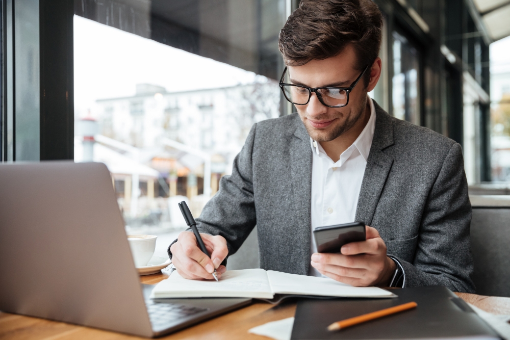 Empresário sorridente em óculos, sentado junto à mesa no café com o computador portátil enquanto estiver usando o smartphone e escrevendo algo para ilustrar assim artigo sobre Dicas para otimizar a gestão financeira de sua empresa com CFO as a Service