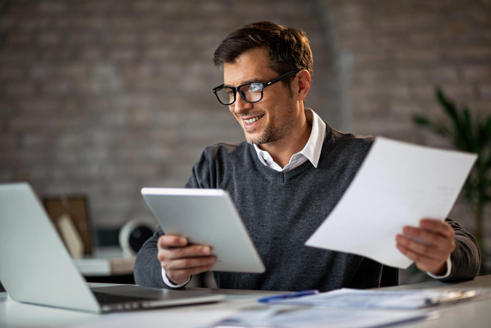 Foto de feliz empresário usando touchpad e laptop enquanto trabalhava em relatórios de negócios no escritório para ilustrar assim artigo sobre CFO as a Service