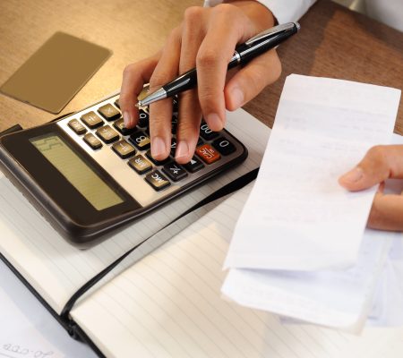 Closeup of person holding bills and calculating them. Notebook, calculator and credit card lying on desk. Payment concept. Cropped view.
